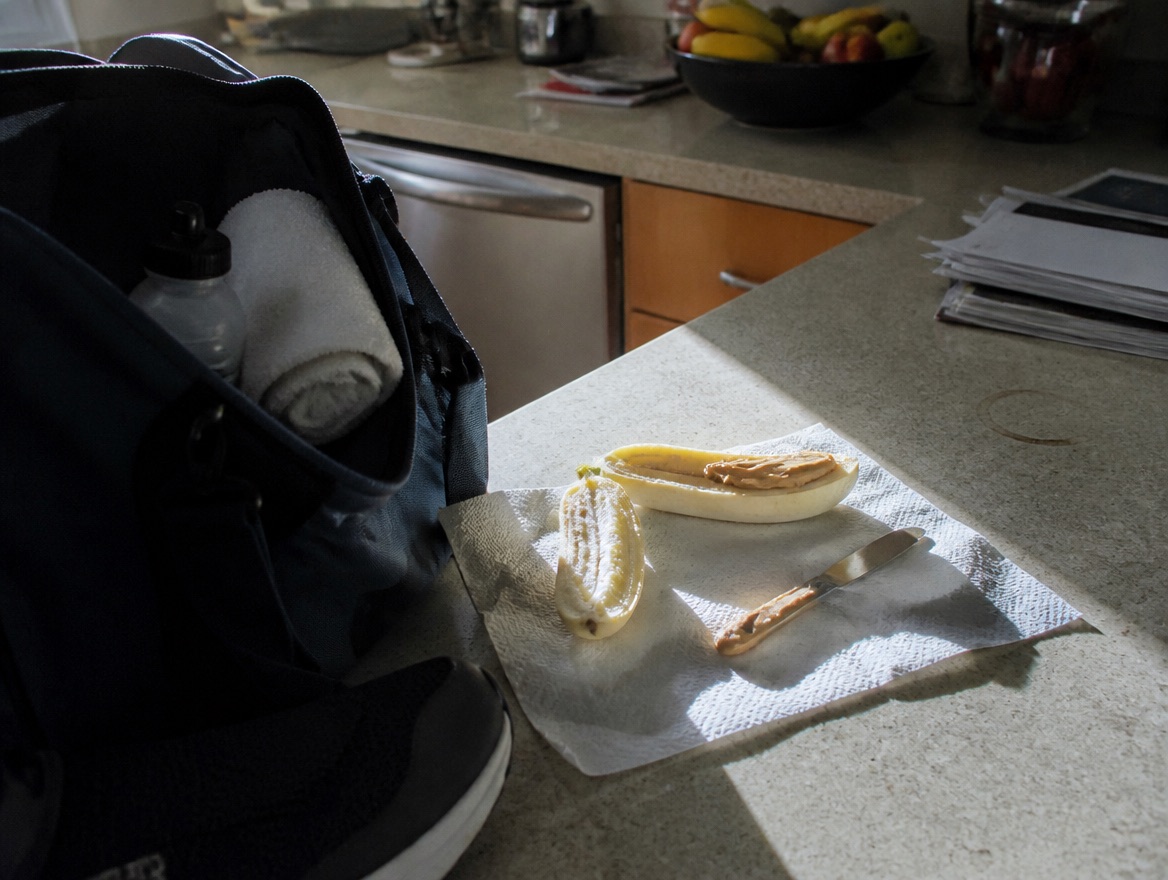 A halved banana spread with peanut butter sits on a paper towel on a kitchen counter beside an open gym bag packed with a towel and water bottle, morning light casting hard shadows across the counter.