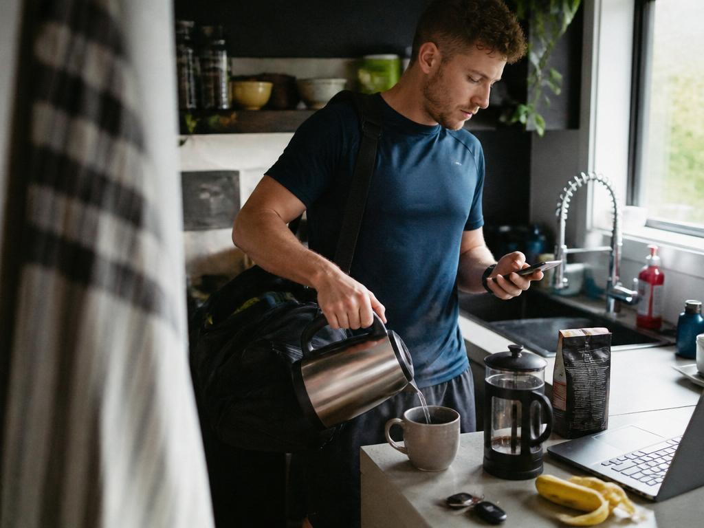 A young man in workout clothes pours hot water into a coffee mug while checking the time on his phone, a gym bag on his shoulder, in a bright kitchen.