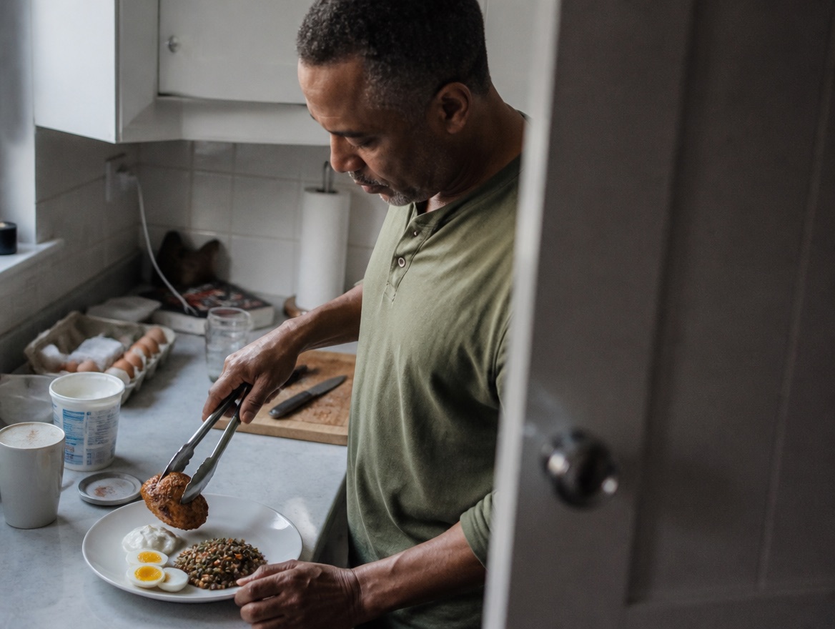 A Black man in his late 40s in an olive henley uses kitchen tongs to lift a piece of grilled chicken onto a plate already holding a halved soft-boiled egg, Greek yogurt and lentil salad, in a softly lit, cluttered home kitchen.